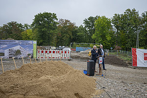 Zwei Männer stehen auf einer Baustelle neben einem Rednerpult und einem Lautsprecher, im Hintergrund Bauschutt, Bauzäune und ein Bauplan auf einer Tafel.