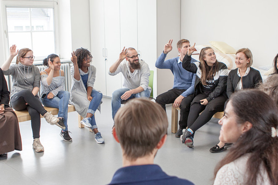Menschen sitzen im Kreis auf Stühlen in einem hellen Raum, einige heben die Hand, andere hören zu.