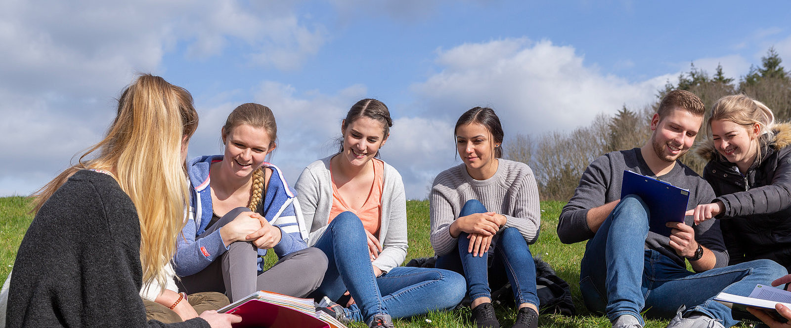 Gruppe von sechs Jugendlichen sitzt im Kreis auf einer Wiese, einige halten Bücher und Notizblöcke, draußen bei bewölktem Himmel