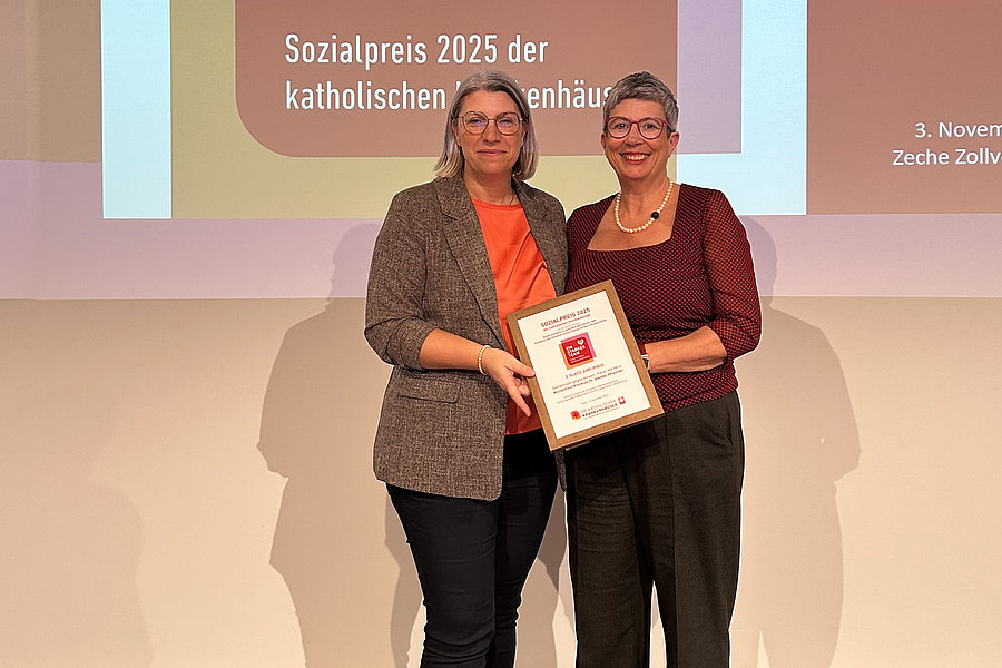 Zwei Frauen stehen nebeneinander und halten gemeinsam eine Urkunde vor einer Projektionswand mit Text