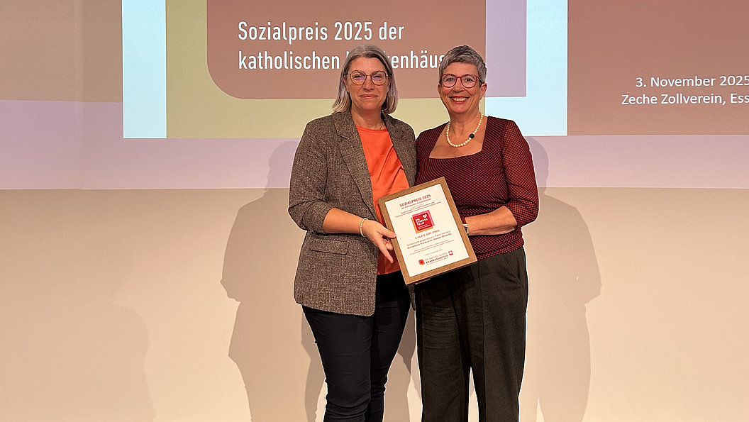 Zwei Frauen stehen nebeneinander und halten gemeinsam eine Urkunde vor einer Projektionswand mit Text