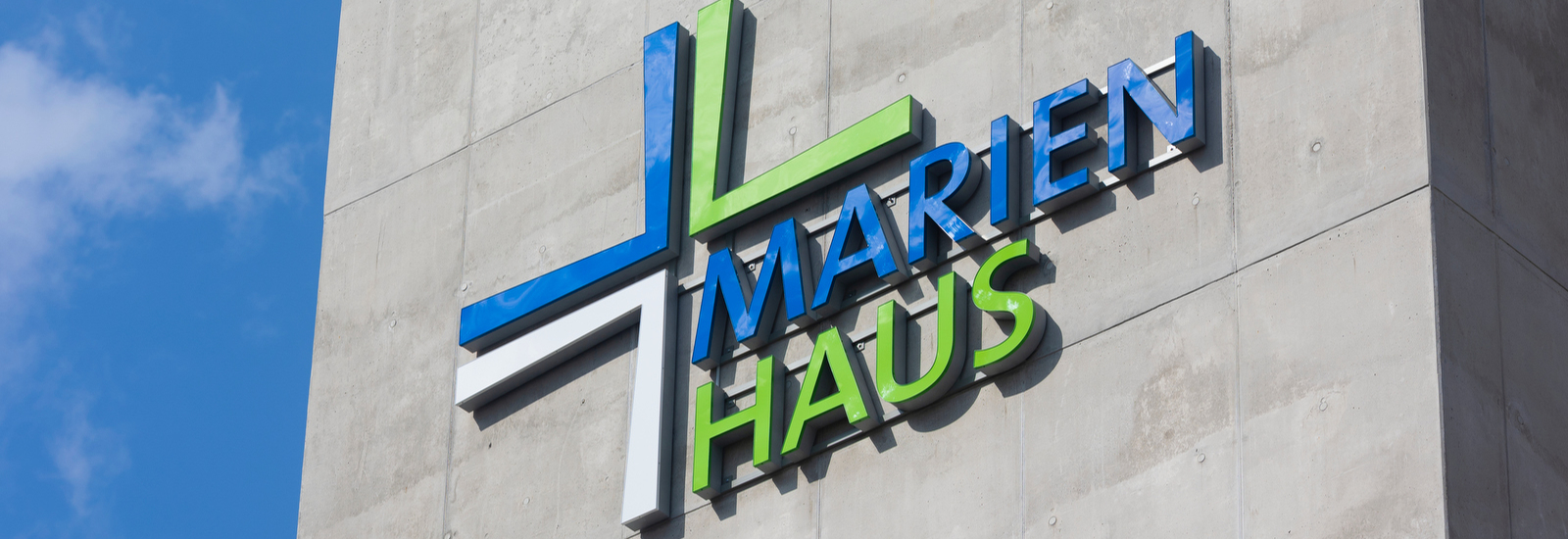 Logo von Marienhaus mit stilisiertem Kreuz aus blauen, weißen, grünen Linien an einer Betonwand