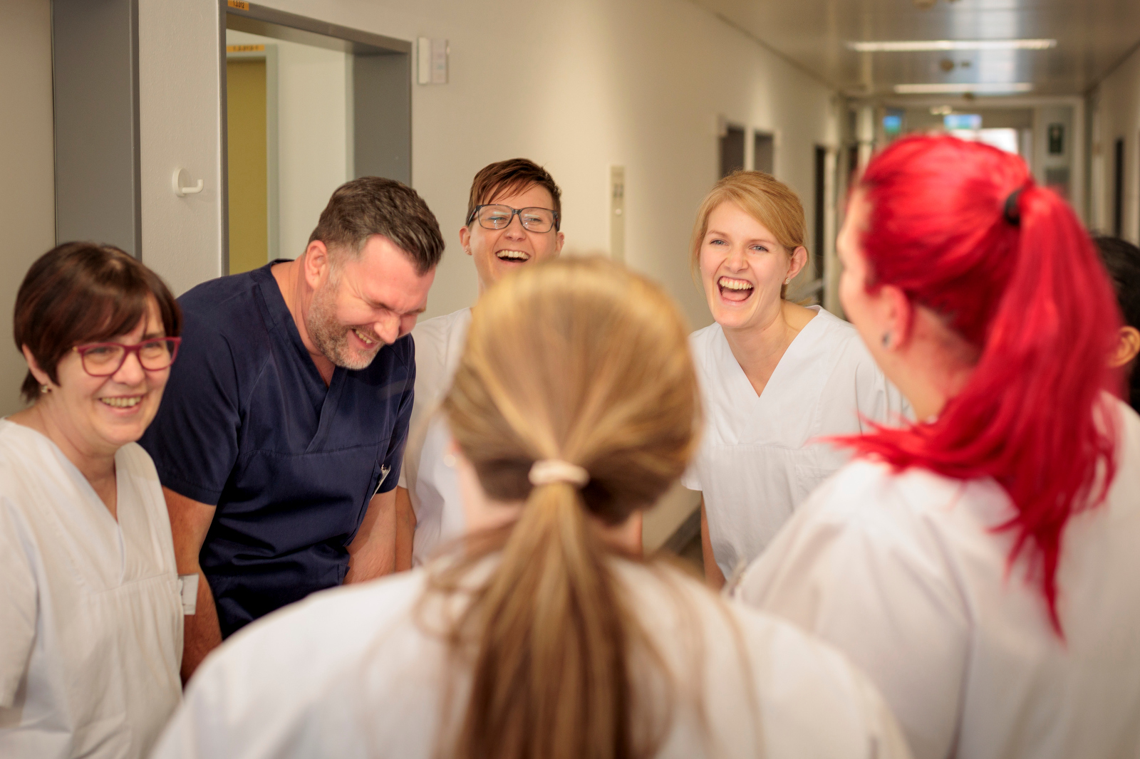 Sechs medizinische Fachkräfte in einem Krankenhausflur lachen gemeinsam