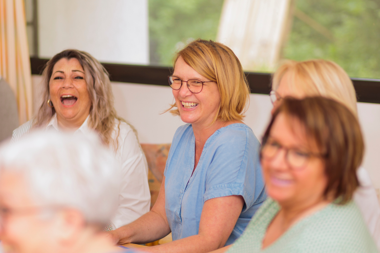 Vier Frauen sitzen zusammen und lachen in einem hellen Raum mit großen Fenstern.