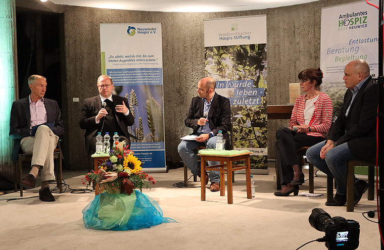 Fünf Personen sitzen auf einer Bühne bei einer Podiumsdiskussion vor Bannern von Hospizorganisationen, eine Person spricht in ein Mikrofon.