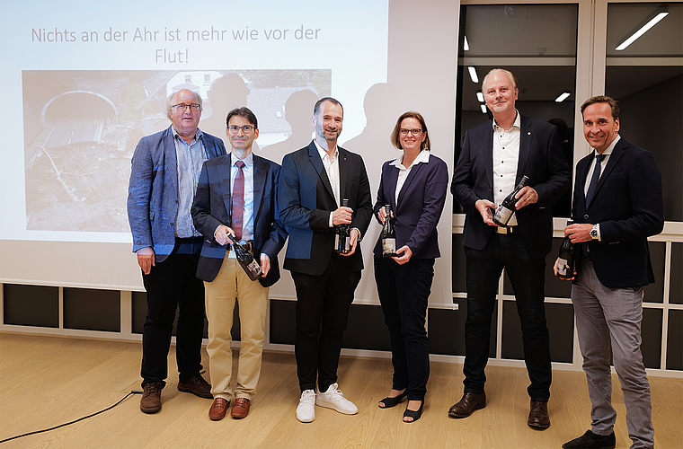 Fünf Personen in formeller Kleidung stehen vor einer Präsentationsleinwand mit dem Text 'Nichts an der Ahr ist mehr wie vor der Flut!' und halten Weinflaschen in der Hand.