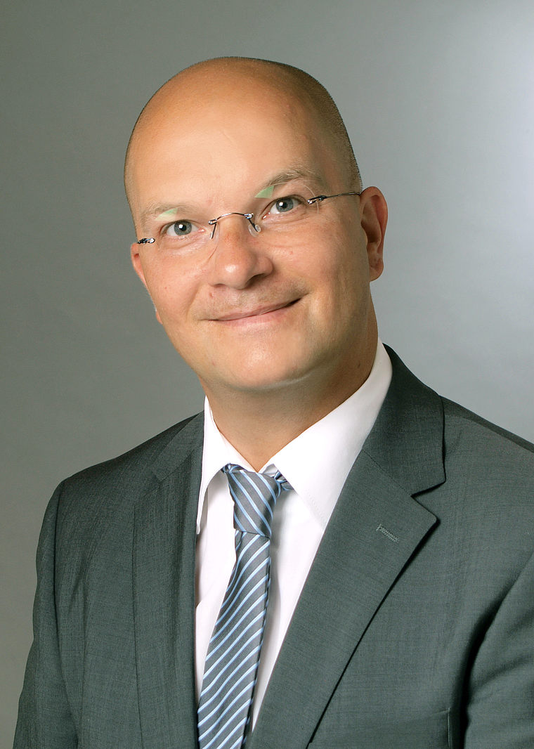 Porträt eines lächelnden Sascha Koenigshoven mit Brille, grauem Anzug, weißem Hemd und gestreifter Krawatte vor neutralem Hintergrund