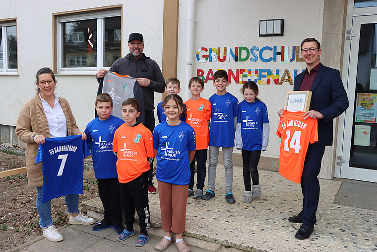 Gruppe von Kindern und Erwachsenen vor der Grundschule Bannflehen mit buntem Schriftzug, einige halten bunte Sporttrikots mit Nummern