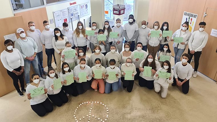 Gruppenfoto von 29 Personen mit weißen Masken und weißen Oberteilen, die grüne Schilder mit Buchstaben halten, die zusammen das Wort 'FREIHEIT' bilden, vor einem Friedenszeichen aus Kerzen auf dem Boden