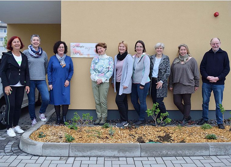 Neun Personen stehen vor einer gelben Wand mit einem Schild 'Hospiz SAPV', sie tragen lässige Kleidung und lächeln