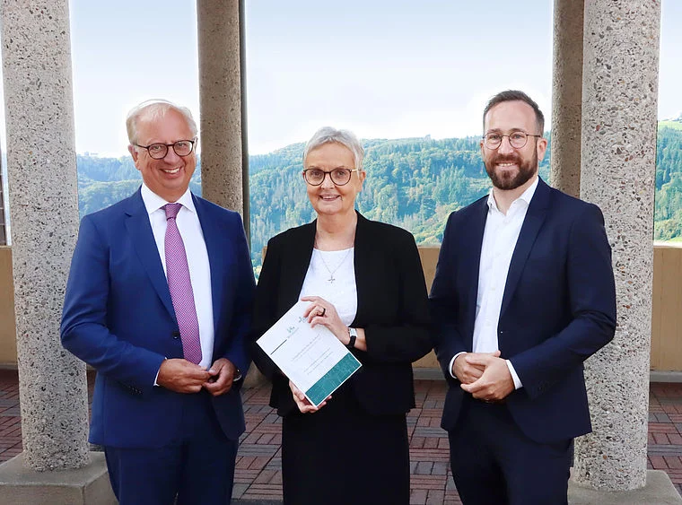 Drei Personen in formeller Kleidung stehen vor Säulen mit Waldlandschaft im Hintergrund, die mittlere Person hält ein Dokument