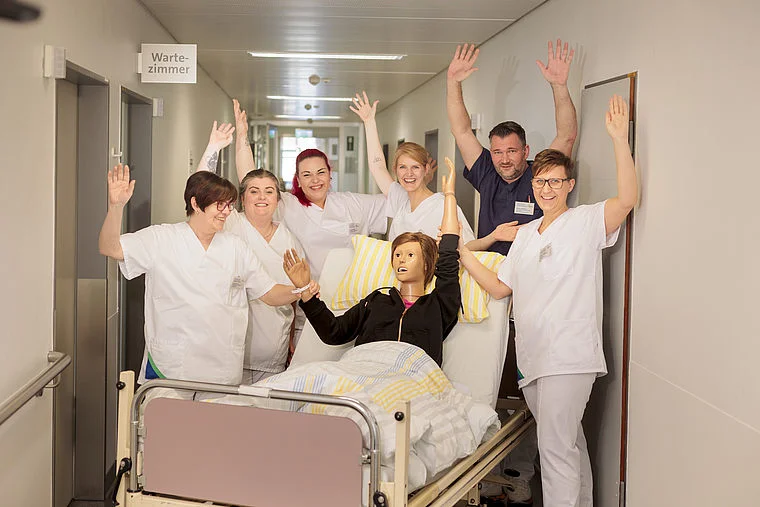 Sechs medizinische Fachkräfte in weißen Kitteln und ein Patient auf einem Krankenhausbett heben die Hände in einem Flur mit Schild 'Wartezimmer'