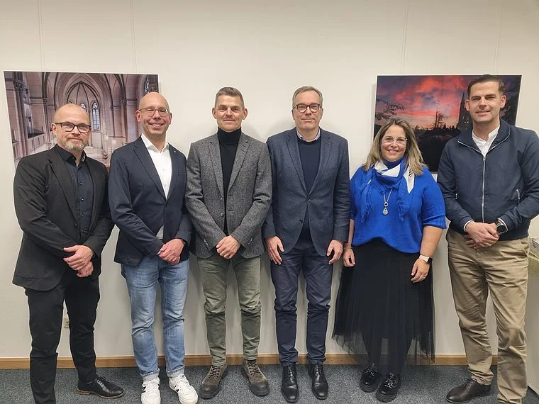 Sechs Personen des medizinischen Beirats stehen nebeneinander vor einer Wand mit zwei großen Bildern, formell gekleidet, in einem Innenraum.