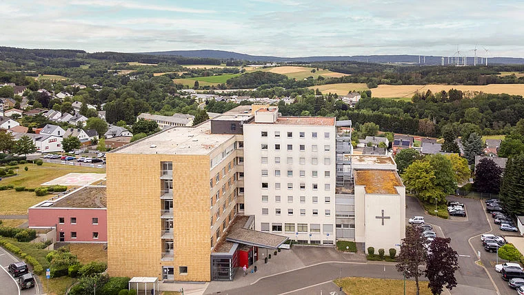 Krankenhausgebäude mit Kreuz auf der Fassade, umgeben von Bäumen und Wohnhäusern in einer ländlichen Gegend