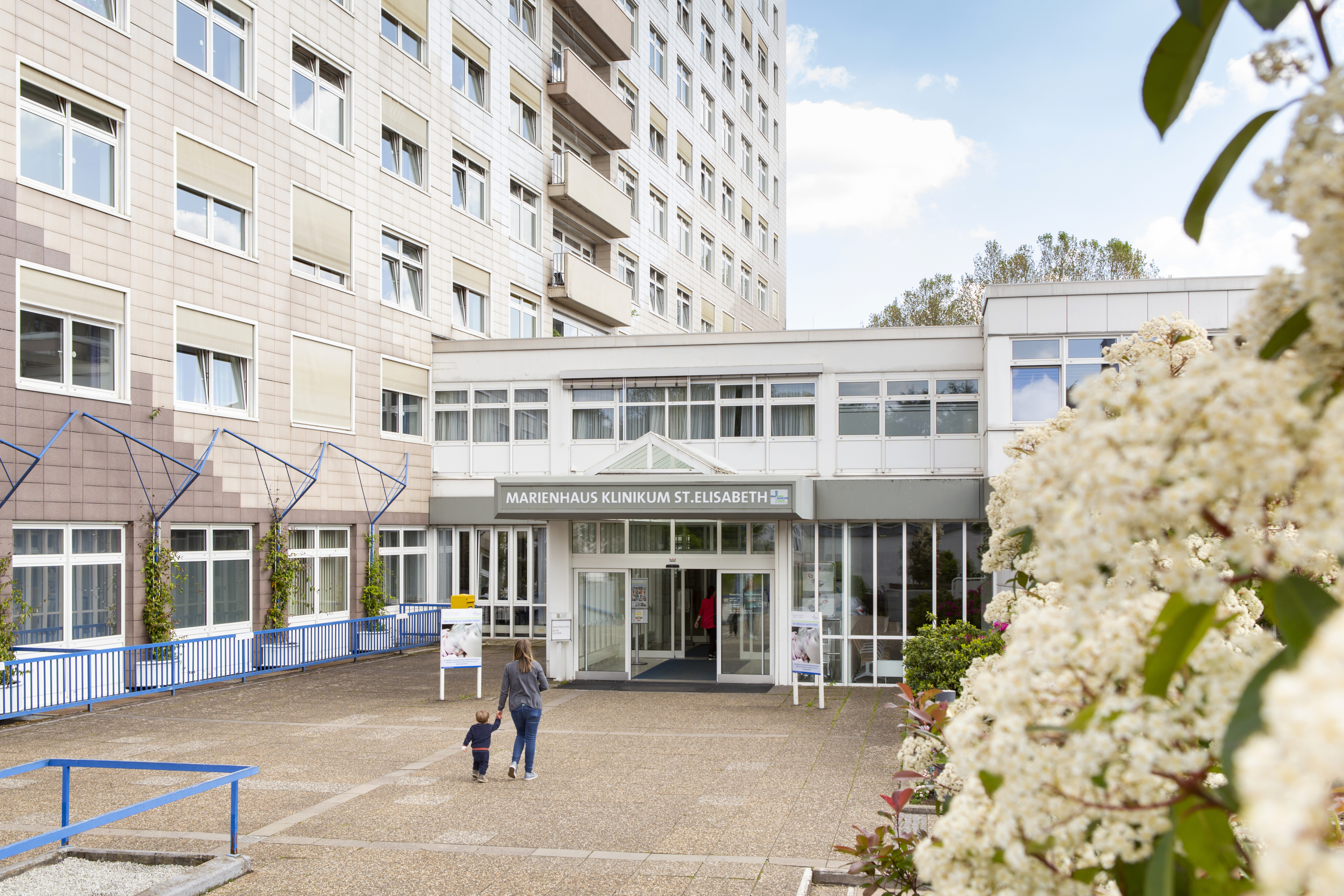 Eingang des Marienhaus Klinikums St. Elisabeth mit einer Frau und einem Kind, die darauf zugehen, umgeben von einem mehrstöckigen Gebäude und blühenden Büschen