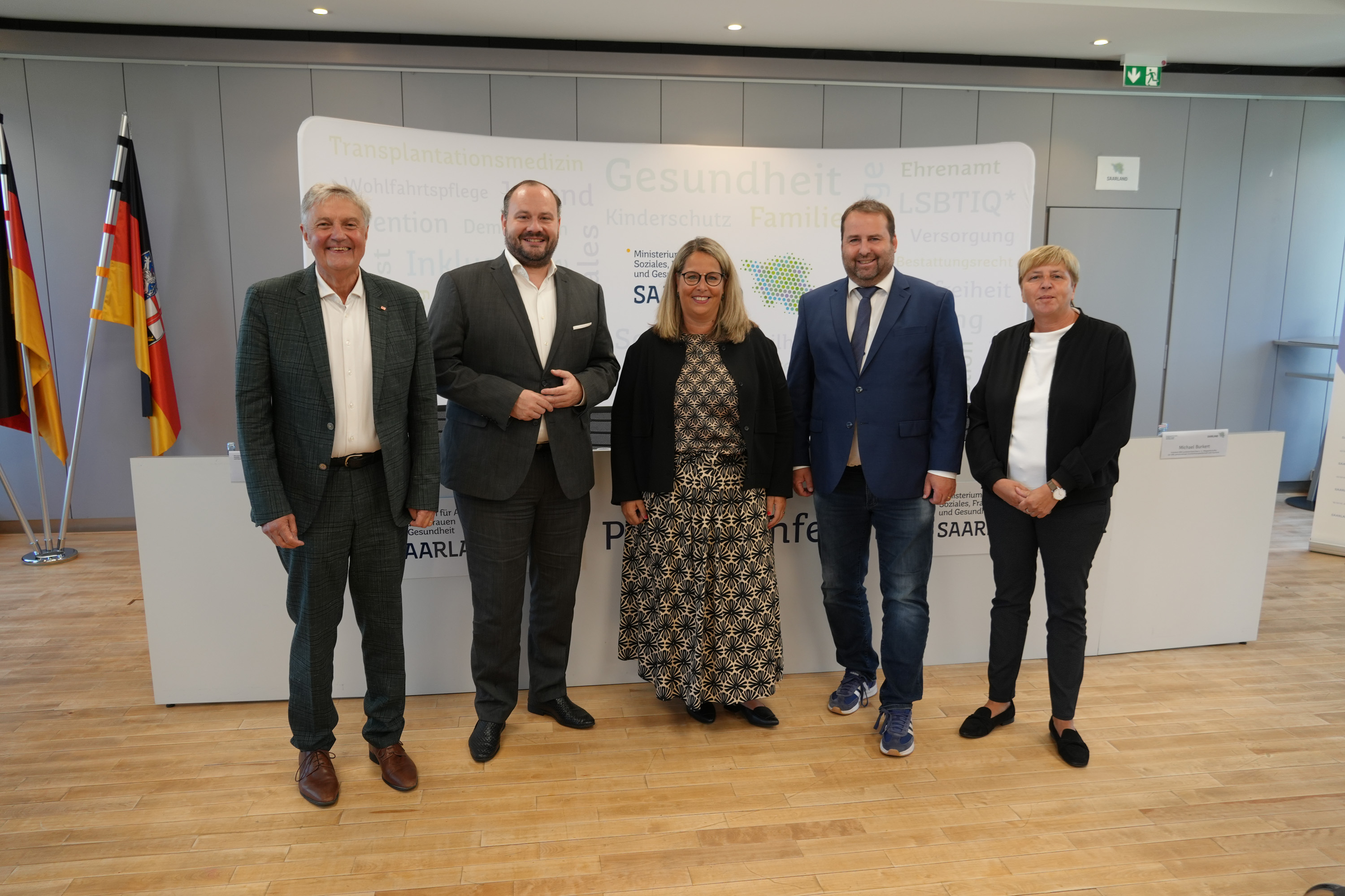 Fünf Personen stehen nebeneinander in einem modernen Raum mit Holzboden und einer weißen Wand im Hintergrund, auf der mehrere deutsche und saarländische Flaggen zu sehen sind