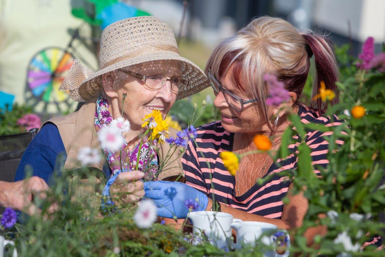 Ältere Frau mit Sonnenhut und jüngere Frau mit gestreiftem Shirt pflücken gemeinsam bunte Blumen im Garten