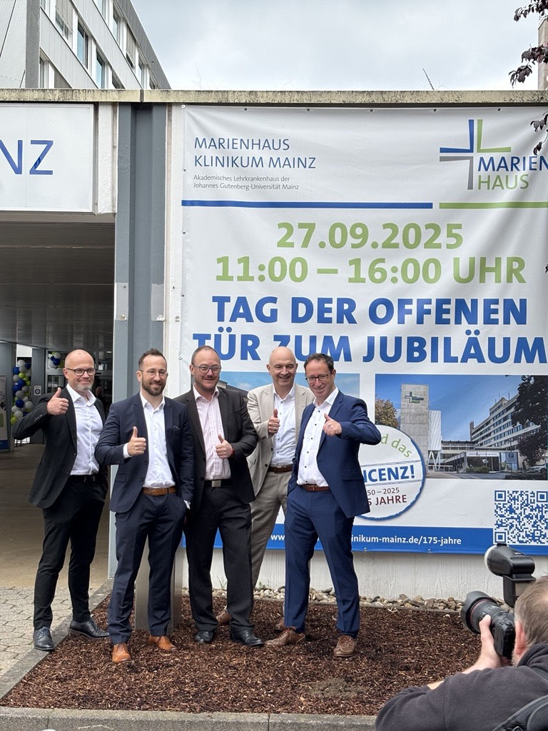 Vier Männer in Anzügen posieren vor einem Plakat mit der Aufschrift '27.09.2025 11:00 – 16:00 UHR TAG DER OFFENEN TÜR ZUM JUBILÄUM' am Marienhaus Klinikum Mainz.
